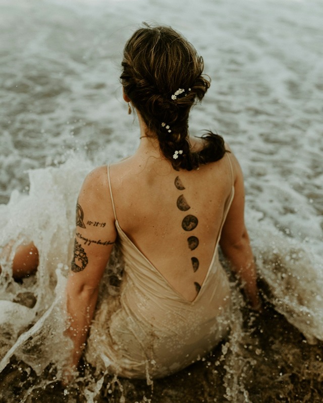 Woman with tattoos of moon on her back sitting in water while waves crash.